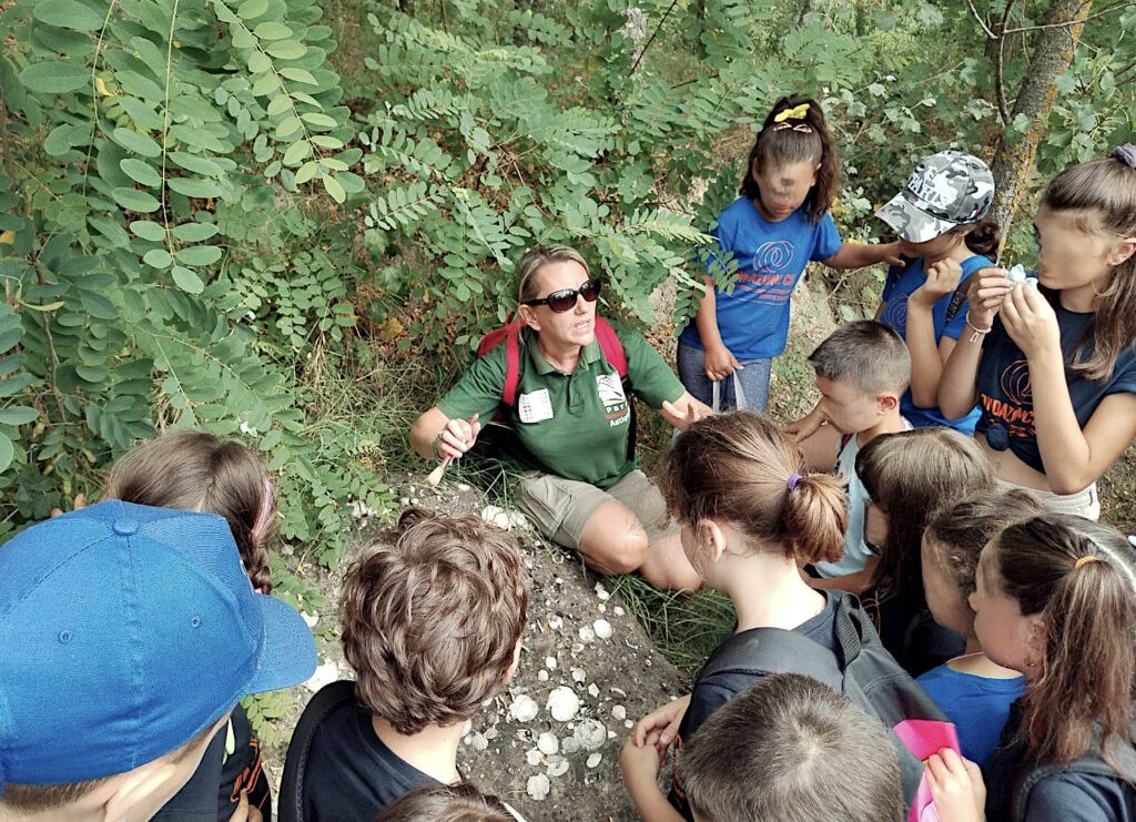 La nostra paleontologa Alessandra, mostra un affioramento di fossili ad una scuola. la sponda indicata è sabbiosa e piena di affioramenti bianchi, poco più su il verde delle felci ricopre la sponda