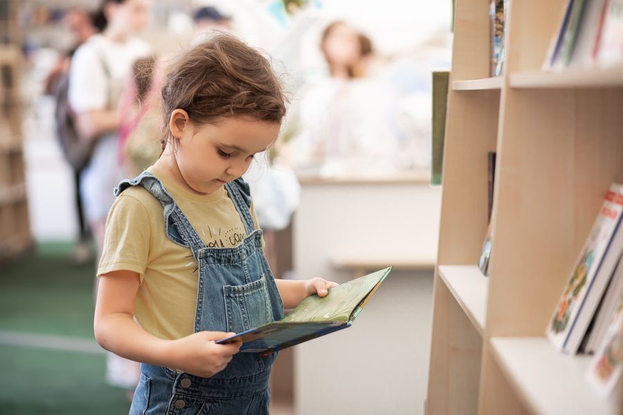 Una bambina sfoglia un albo illustrato davanti agli scaffali di una biblioteca